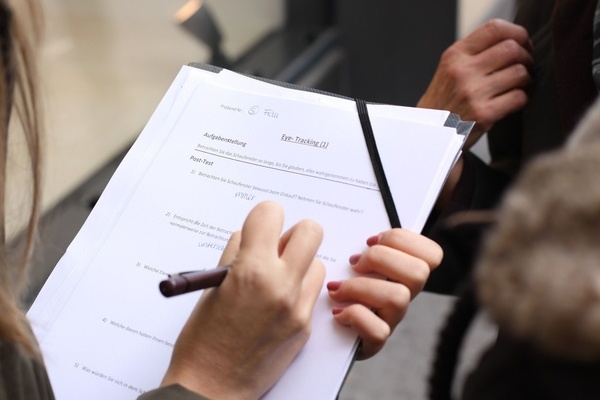 Eine Person mit langen Haaren mit Stift in der Hand füllt ein Formular auf einem Klemmbrett aus- gegenüber steht eine Person, die von ihr befragt wird